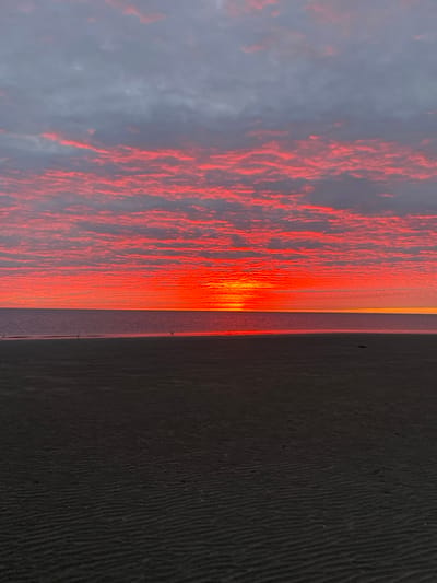 A horizontal, full-frame shot captures a sunset over the White Sea in Severodvinsk, Russia. The sky is dominated by a vibrant orange and red glow from the setting sun, which is visible as a bright band on the horizon. The clouds are tinged with pink and orange, contrasting with the darker gray clouds above. The sea is calm and reflects the colors of the sky. The foreground features a dark, sandy beach with subtle rippled textures. A few small birds are visible on the wet sand near the waterline. The overall impression is one of a serene and colorful natural landscape.