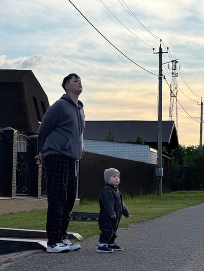 Two individuals are visible. An older person wears a grey hoodie and checkered pants, and white sneakers. A younger person wears a dark jacket, dark pants, a light-colored beanie, and dark sneakers. Both are looking upwards at the sky in an outside setting. The location appears to be in the city of Vysokinichi, Russia. A utility pole and power lines are in the sky. Houses and greenery are in the background.