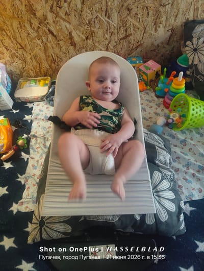 A baby is sitting in a white baby bouncer. The baby is wearing a camouflage tank top and a light-colored diaper. The baby is looking at the camera and smiling. There are toys scattered around the baby bouncer. In the background, there is a wall made of OSB boards. The floor is covered with a patterned rug. The text overlay at the bottom indicates the photo was taken on a OnePlus 1 with Hasselblad, in Pugachyov, Russia, on June 21, 2026, at 15:36.