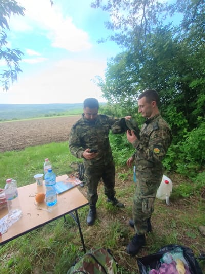 Two men in camouflage uniforms stand near a folding table set up in a grassy area. The table holds water bottles, an orange, a cup, and food wrappers. A field is visible in the background, along with trees and a blue sky with clouds. A black trash bag and a white canister sit on the ground near the men. The man on the left adjusts the other man's cap. The location is near Kostena Reka, Bulgaria, based on the landscape.