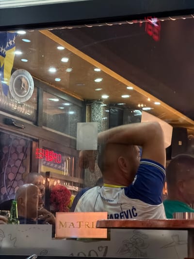 The image shows a reflection in a glass window, possibly of a shop or cafe, in Nević Polje, Bosnia and Herzegovina. Inside, people are visible, some appearing to be patrons. A clock on the wall indicates a time between 11:50 and 12:00. A red neon sign is illuminated. A Bosnian flag is partially visible. The foreground shows a table with what appears to be a beer bottle and glasses. The overall lighting suggests it is evening or nighttime.