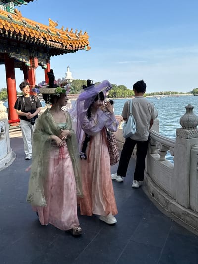 An outdoor scene depicts a group of people enjoying a sunny day at a historic Chinese park or garden, likely in Xicheng District, China. The blue sky is clear above a large body of water, possibly a lake, lined with distant green trees and a white pagoda on the horizon.
In the foreground, an ornate traditional Chinese architectural structure, featuring a golden and red tiled roof with intricate carvings, frames the left side of the image. Along the right, a stone balustrade overlooks the water.
Four main individuals are visible. Two women are dressed in elaborate traditional Chinese attire (Hanfu). One wears a flowing green and pink dress with a broad-brimmed hat adorned with flowers, while the other is in a purple and pink ensemble with a sheer purple veil. Both have intricate hairstyles. They appear to be posing or simply standing, enjoying the setting.
Behind them, a man in a light beige shirt, dark pants, and white sneakers stands by the balustrade, looking out at the water while holding a smartphone. He carries a light blue shoulder bag. To the left of the women in traditional dress, a man wearing glasses, a black t-shirt, and white pants looks towards them, holding what appears to be a camera or phone. The text "LUMIX S5" is visible on his t-shirt, identifying the camera brand.
The overall atmosphere is leisurely, suggesting a tourist outing, cultural event, or a photoshoot. The lighting indicates it is daytime, with clear weather.