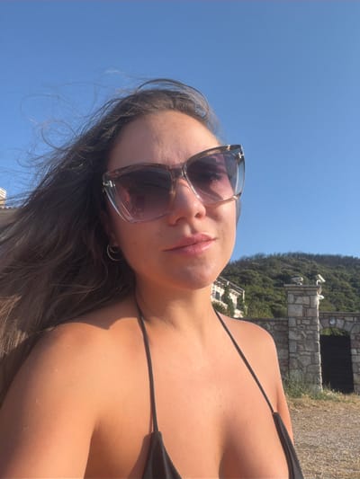 A young woman with long, dark, wavy hair is captured in a selfie with a clear blue sky in the background. She is wearing large, translucent sunglasses with a subtle tortoise shell pattern and a black bikini top. Her hair is windblown, framing her face. The background shows a hilly landscape with green vegetation and a stone structure, possibly part of a building or an old gate. In the distance, two security cameras are visible mounted on a stone pillar. The scene appears to be outdoors, under bright sunlight. The location is identified as Buljarica, Montenegro, suggesting a coastal or hillside setting.
