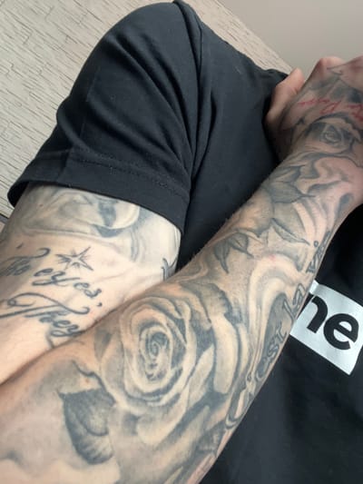 The image shows a person's arms and part of their torso. The person is wearing a black t-shirt with a white box logo containing the letters "ne". The arms are heavily tattooed with black and grey designs, including roses, flowers, and lettering. The person is sitting down, with one arm resting on the other. The background is blurry, but suggests a simple interior space. There is no indication of the image being taken in Belovo, Bulgaria.