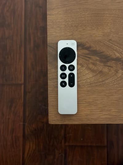 The image shows a white Apple TV remote lying on a light brown, wood-grained surface. The surface appears to be part of a table or similar piece of furniture. In the lower right of the frame, darker brown, wood-like flooring is visible. There are no people or specific identifying features that could link this scene to Buford, United States. The subject matter is a static shot of a remote control and furniture.