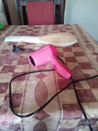 A pink hairdryer with a black cord and plug lies on a table covered with a checkered tablecloth. A loofah brush is placed next to the hairdryer, with its white fabric end on the left and the rough, natural side on the right. The background shows a portion of a wooden chair with a red seat. The scene is indoors, likely in a domestic setting in the city of Luka, Ukraine.
