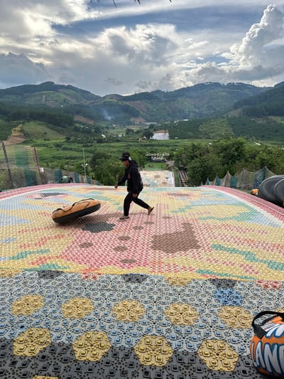 In a scenic landscape at Thôn 5, Vietnam, a person is captured mid-stride on a vibrant, multi-colored patterned walkway. The walkway is composed of interlocking hexagonal tiles, featuring a variety of colors including yellow, red, green, and blue, interspersed with a grid-like pattern of small dots and squares. To the right of the walkway, a black tube-like object with a tan strap lies on the ground. The person, wearing a black hoodie, black pants, and a black baseball cap, appears to be walking away from the camera towards the right side of the frame.
The background reveals a lush, mountainous terrain under a partly cloudy sky. The mountains are covered in dense green vegetation, suggesting a tropical or subtropical environment. Rolling hills descend into a valley where a small settlement or resort is visible, with buildings scattered among the trees. In the distance, a sign is partially visible, with text that appears to read "MONGOLAND DALAT". The lighting suggests it is daytime, possibly late afternoon, given the soft light and the shadows cast by the person. The clouds are a mix of white and grey, indicating dynamic weather conditions. The overall scene evokes a sense of outdoor recreation and exploration in a beautiful natural setting.