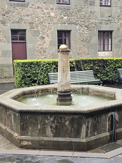 The image shows a stone fountain in Алупкинский городской совет, Ukraine, set against a building with stone walls and windows with dark frames. A green hedge and a green bench are located behind the fountain. Water streams from the central pillar of the fountain into a stone basin filled with water. The fountain's structure shows signs of weathering and minor damage. The ground around the fountain is paved with stone tiles.