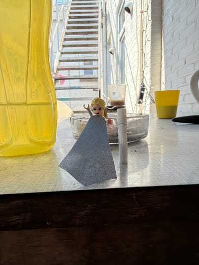An Elsa doll with a light blue cape stands on a textured white table in Boxtel, Netherlands. The doll is positioned in front of a clear glass ashtray containing cigarette butts and ash. To the left of the doll is a large, translucent yellow bottle, possibly containing liquid. In the background, an outdoor metal staircase with visible steps and railings ascends. Beyond the staircase, a brick building with windows is visible, along with some string lights. A yellow cup is also present on the table to the right of the ashtray. The scene is illuminated by natural light, casting shadows on the table.