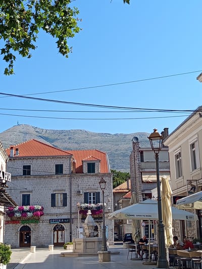 The image captures a sunlit street scene in Trebinje, Bosnia and Herzegovina, likely during the daytime. In the foreground, a cobblestone street leads towards a square with outdoor seating for a cafe, complete with tables and umbrellas. Several people are seated at the tables, appearing to be patrons of the establishment.
The architecture suggests a Mediterranean influence, with buildings constructed from stone and featuring terracotta tiled roofs. Some buildings have flower boxes adorning their windows, adding a touch of color to the scene. Street lamps and a decorative fountain are also visible, contributing to the ambiance of the public space.
In the background, a large hill or mountain range dominates the skyline, with a telecommunications tower visible near its summit. The sky is clear and blue, indicating pleasant weather conditions. Power lines are strung across the upper portion of the image, a common sight in urban environments.
The overall impression is one of a tranquil, European town square on a clear day, with a blend of historical architecture and modern elements.
