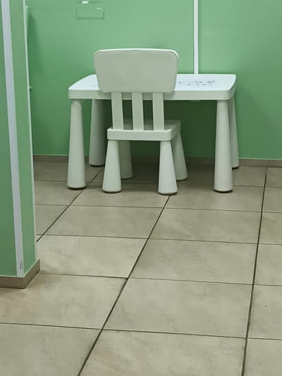 A white child-sized table and chair are positioned in a room with light green walls and a beige tiled floor. The table has four conical legs, and the chair has a backrest with vertical slats and a seat. The table surface has a faint outline of what appears to be a cartoon character. The scene is well-lit, and there are no people or animals present. The image appears to be taken from a medium height, looking slightly downwards towards the floor. The overall environment suggests a children's play area or classroom.