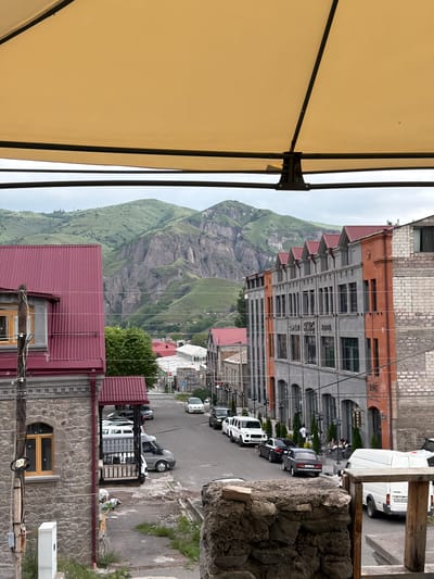 A street view in Goris, Armenia, is captured from an elevated position, possibly a balcony or a higher floor, looking down a road lined with buildings and a backdrop of green, rocky mountains. The foreground is partially obscured by a large, beige awning.

On the left, a building with a dark red, sloped metal roof and stone exterior features a single, arched window with a wooden frame. A wooden pole with electrical wires is positioned in front of this building.

The street is paved and has parked cars along its sides. Vehicles visible include a white Mercedes-Benz G-Class SUV, several passenger cars, and white vans. Some people are seen walking on the sidewalk in front of a hotel.

Dominating the mid-ground is a multi-story building with a grey facade and orange accents. It has many arched windows and signage that reads "BMG HOTEL". Further down the street, other buildings with red roofs and varied architectural styles are visible, suggesting a densely populated urban area.

In the background, impressive, steep, green mountains with visible rock formations rise against a slightly overcast sky. The overall impression is a blend of urban infrastructure and natural mountainous landscape.