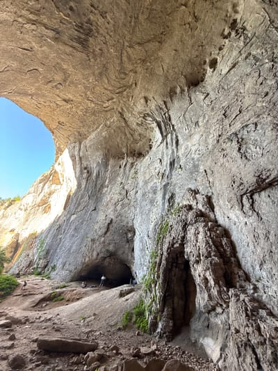 This image depicts the interior of a large natural cave in Karlukovo, Bulgaria. The cave's ceiling is a massive, rough rock formation with visible striations and some circular depressions. The walls are also composed of textured rock, with one section on the right featuring a prominent, layered rock formation covered in moss and greenery, resembling a smaller, rough-hewn archway or opening.
In the foreground, the ground is uneven and covered with rocks and dirt. Several people are visible throughout the scene. Closer to the cave entrance, a small group of individuals is seated or lounging, with some equipment scattered around them. Further into the cave, a single person stands near the mouth of a darker cavern. On the left side of the image, on the exterior slope leading up to the cave, a few more individuals are scattered, one walking and others appearing to be seated. The bright blue sky is visible through a large opening on the left, indicating the cave is open to the outside. Sunlight streams in, illuminating the left-hand rock face. The overall impression is of a natural geological formation being explored by visitors.