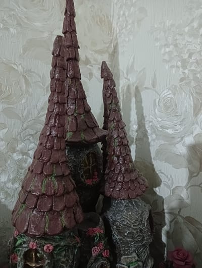 The image shows a close-up of three decorative, cone-shaped structures resembling miniature fairy houses or whimsical towers. The structures have a textured, dark reddish-brown roof made of overlapping scales, with subtle green accents suggesting moss or vines. The bases of these structures are dark gray and rough, mimicking rock or tree trunks, and are adorned with small pink artificial roses and green foliage. The background is a textured wallpaper with a light, floral pattern of roses and leaves, creating a soft, slightly out-of-focus backdrop. The lighting suggests an indoor environment, possibly a shelf or display area. No people are visible in the image. The location in Bender, Moldova is not discernible from the visual information provided in the image.