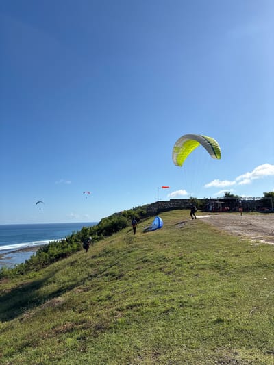 The image depicts a paragliding launch site in Benoa, Indonesia, with a green and yellow paraglider inflating near the edge of a grassy cliff overlooking the ocean. A person is preparing for takeoff.  Other paragliders are visible in the sky above the ocean, and people are observing near the top of the cliff. A small orange windsock is positioned on the cliff's edge. Vegetation lines the coastline.
