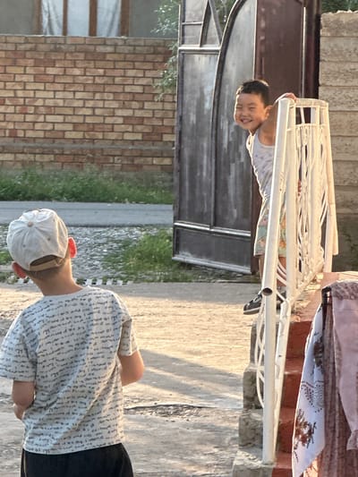 This media file depicts two young boys in an outdoor setting, possibly a yard or backyard, in Тайгараев, Kyrgyzstan. The scene appears to be bathed in warm, late afternoon or early evening sunlight, as indicated by the long shadows and golden hue.
The boy in the foreground is wearing a white baseball cap and a light-colored shirt with a repeating pattern of small text or symbols. He is facing away from the camera, with his arms bent at the elbows and his hands clasped behind his back. His posture suggests a relaxed or perhaps curious stance.
In the background, a second, younger boy is visible, leaning over a white metal structure that resembles a drying rack or a playground element. He is wearing a light-colored tank top with a subtle pattern. His head is tilted back, and he is smiling broadly, with his mouth open in what appears to be laughter. His joy and carefree expression are a notable detail.
The setting includes elements of a residential area: a brick wall on the left, a metal gate or door, and a paved pathway. The presence of laundry on the white structure in the background suggests it might be a clothes drying area. The overall atmosphere is one of casual outdoor play and familial warmth.