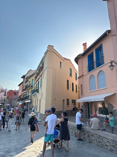 The image depicts a sunlit cobblestone street in Collioure, France, lined with colorful buildings. In the foreground, several people are walking, including a man in turquoise shorts holding a child, a woman in a black and white polka dot dress, and a man in a white t-shirt and black shorts. To the right, a pink building with blue shutters stands prominently. Further down the street, more people are seen browsing shops and walking along the waterfront. The sky is a clear, pale blue, with a single bird visible in the upper left quadrant. The architecture suggests a Mediterranean coastal town.