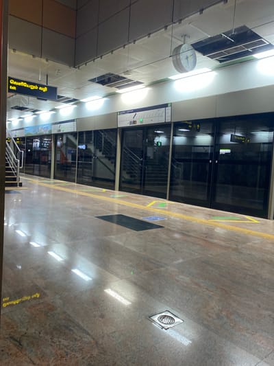 The image shows an empty platform at a metro station in Chennai, India. The platform is made of polished granite-like tiles, with a tactile paving strip running along the edge. There are several doors on the right side of the image, which appear to be the entrances to the metro train. Above the doors, there are signs in Tamil and English, indicating "Exit". A clock is visible hanging from the ceiling. The lighting is artificial, with fluorescent lights reflecting off the shiny floor. There are no people or trains visible in the image, suggesting that the metro station is either closed or the photo was taken during off-peak hours. The overall impression is one of modern infrastructure, clean and well-maintained.