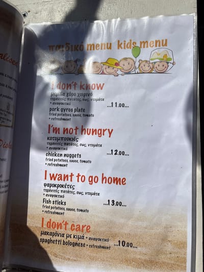 The image displays a laminated children's menu from a restaurant located in the city of Saint Arsenius, Greece. The menu's top section features the title "παιδικό menu kids menu" in orange and green text, along with an illustration of five cartoon children's faces peeking over a horizontal line. One child wears a yellow hat, another holds a green balloon, and a third wears a hat depicting a building with mountains.

Four menu items are presented with humorous English titles in red font: "I don't know" (pork gyros plate), "I'm not hungry" (chicken nuggets), "I want to go home" (fish sticks), and "I don't care" (spaghetti bolognese). Each item includes supplementary details in black text: the first three list "fried potatoes, sauce, tomato + refreshment," while the last lists "+ refreshment." Corresponding prices of 11.00, 12.00, 13.00, and 10.00 are listed next to each item. The bottom portion of the menu features a printed sandy texture as a background element.

The menu is positioned outdoors under direct sunlight, which creates significant glare across its laminated surface, particularly in the central and upper-right areas. Dark, angular shadows, likely from an overhead object or person, are cast prominently across the menu, obscuring parts of the text and illustrations on the left side. The menu exhibits signs of use, including minor creases and smudges. A partial view of an adjacent, bound menu page is visible along the left edge of the frame.