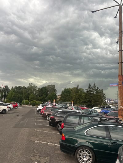 The image shows a parking lot filled with cars under a cloudy sky. The cars are parked in rows on an asphalt surface marked with white lines. In the foreground, a dark green BMW station wagon is visible, featuring a roof rack and silver alloy wheels. Several other cars, including a blue hatchback, a red hatchback, and white cars, are also present.
Behind the cars, a line of trees with lush green foliage separates the parking lot from a few buildings. A sign for "MRÓWKA" is visible on one of the buildings, suggesting it might be a retail store. The sky is filled with dramatic, dark gray clouds, indicating overcast weather, possibly before or during a storm. The overall lighting suggests it is daytime, though the cloud cover makes it appear dim.
The image is oriented vertically, with the sky occupying the left half and the cars and parking lot on the right. A wooden structure with what appears to be lighting fixtures runs along the top edge of the image. There are no people clearly visible in the frame, and the overall mood is somber due to the weather. The location is identified as Jastrzębie-Zdrój, Poland.