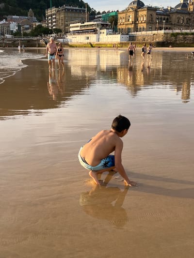 This is a vertically oriented, wide-angle photograph taken on a sandy beach in San Sebastián, Spain. The image features several people scattered across the beach and shallow water, with buildings and lush green hills visible in the background. A young boy in blue patterned swim trunks is the most prominent subject, sitting with his back to the camera on the wet sand. He is leaning forward, with his hands resting on the sand as if preparing to push himself up. His shadow, elongated by the low angle of the sun, stretches out before him.

Further down the beach, a group of people are wading in the shallow water, their reflections mirrored on the smooth, wet sand. They appear to be enjoying a leisurely day at the beach. The lighting suggests it is either early morning or late afternoon, with the sun casting long shadows and creating a warm, golden hue across the scene. The water is calm and reflective, adding to the serene atmosphere. The architectural style of the buildings in the background, with their ornate facades and balconies, hints at a European coastal city. There is no visible text in the image.