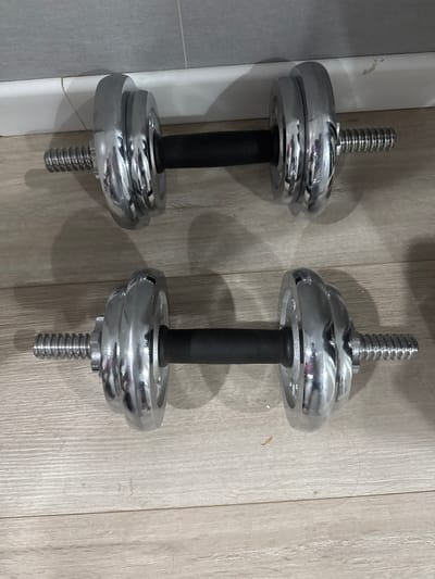 Two chrome dumbbells with black grip handles lie parallel on a light-colored wooden floor. Each dumbbell has threaded ends with plates attached. The dumbbells are positioned horizontally. In the background, a white baseboard runs along a gray tiled wall. The scene appears to be indoors, possibly a home gym or storage area. There is no indication of the location being in Zaşağan, Kazakhstan.