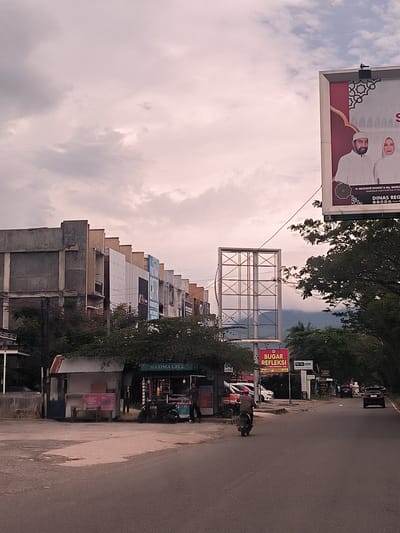 This image captures a street scene in Lampeuneurutgampong, Indonesia, under an overcast sky that gives the light a soft, diffuse quality. The foreground features an asphalt road with a person on a motorcycle driving away from the viewer. To the left, a small, shed-like stall is visible, shaded by trees, with the text "HAZIMA CELL" above its entrance. Another small stall with a pink sign stands next to it, and a man is seen standing near the HAZIMA CELL stall.

In the midground, a row of commercial or residential buildings lines the left side of the street, some appearing weathered or unfinished with exposed concrete, while others have more modern cladding. A large, empty metal framework for a billboard is prominent, with distant mountains faintly visible behind it through the haze. On the right, a large billboard dominates the upper portion of the frame, displaying an advertisement featuring two people identified as "H. MUZAKIR MANAF & NY. MARLIAN," with accompanying text "GUBERNUR ACEH" and "DINAS REG." The billboard is adorned with Islamic geometric patterns.

Additional street signs include a red rectangular sign stating "BUGAR REFLEKSI" and a smaller "METAL SPORT" sign. Trees with dense foliage are interspersed throughout the scene. The overall impression is one of a busy, yet typical, urban or suburban thoroughfare in the region.