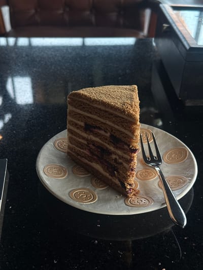 A forensic description of the scene in Tibubeneng, Indonesia, reveals a close-up, eye-level shot of a single slice of multi-layered cake positioned on a decorative plate, accompanied by a fork, resting on a dark, reflective table surface.

The cake slice stands upright, presenting approximately ten to twelve thin, brown cake layers alternating with lighter cream-colored frosting. Interspersed within some of the cream layers are streaks of dark, possibly berry-based, filling. The cake's top and visible side are covered in fine, light-brown crumbs.

The cake rests on an off-white or light gray circular ceramic plate, which features multiple raised, circular, light-brown decorative patterns resembling medallions or cookies. To the right of the cake on the plate lies a silver-colored metal fork, with its tines oriented towards the cake.

The plate and fork are situated on a dark, polished, granular surface, likely a granite countertop or table, which exhibits minor light speckles and distinct reflections of light sources and the plate. In the blurred background, a segment of a brown, upholstered piece of furniture (possibly a booth or chair) is visible on the left, and a dark, angular object (possibly a cabinet or display unit) is on the right. Diffuse light sources, likely windows or interior lighting, are reflected in the table's surface. No individuals are visible in the image.