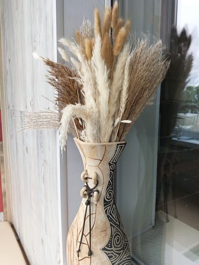 An arrangement of dried grasses and reeds is displayed in an ornate, tall ceramic vase. The vase has a textured, cream-colored finish with intricate black tribal patterns etched into its surface. The dried botanical arrangement is a mix of colors, including light brown, beige, and cream, with various textures from fluffy plumes to slender stalks. The vase and arrangement are positioned in front of a large window, likely in a home or business, with a reflection of the outside environment visible in the glass. The background to the left consists of a wall with a light wood-paneled texture. The setting appears to be indoors, with natural light coming from the window. The image does not provide specific location details within Novyy, Russia.