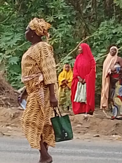 The image captures an outdoor scene featuring several individuals, predominantly women, against a lush green background of foliage.

In the foreground, slightly to the left, a dark-skinned woman walks from left to right, shown in profile. She wears a long, flowing dress and a matching headwrap, both adorned with a distinctive yellow and brown wavy or serpentine pattern. In her right hand, she carries a dark green tote bag, while her left hand holds a light-colored smartphone or similar device. Her bare feet are visible as she walks.

Behind her, in the midground to the right, stands another dark-skinned woman, facing slightly left, with a warm smile on her face. She is dressed in a vibrant, full-length red garment, possibly a hijab or abaya, which includes a head covering. Her hands are clasped in front of her, holding a light grey or white folded cloth or bag.

Further in the background, between the walking woman and the red-dressed woman, a smaller figure, likely a child, is partially visible. This individual wears a bright yellow hooded garment and appears to be standing or sitting. To the far right in the background, another dark-skinned woman stands, wearing a light tan or beige head covering and matching attire. Her lower body is partly covered by a colorful, patterned fabric, and she appears to be holding a small object or bundle in her arms, possibly a baby.

The background consists of dense green foliage, likely bushes or trees with large, lobed leaves, suggesting a tropical or subtropical environment. The ground beneath the individuals appears to be earthy and uneven, with a darker grey strip at the very bottom, which could be a road or pathway. The lighting suggests it is daytime, possibly under soft or diffused sunlight. No text is visible in the image.