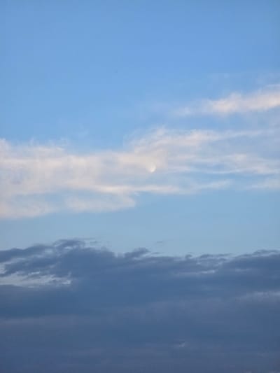 This is a vertical, close-up photograph of the sky. The sky is predominantly a clear blue with some wispy, white clouds. On the left side of the frame, a faint, pale crescent moon is visible, partially obscured by a thin cloud. Towards the right, a band of darker, denser clouds stretches across the frame, creating a distinct contrast with the lighter sky. The clouds appear textured and somewhat layered. The overall impression is of a daytime sky, possibly late afternoon or early evening, given the presence of the moon and the gradation in cloud darkness. There are no discernible people, objects, or text in the image, only the vast expanse of the sky and its cloud formations. The location context is Yevpatoriya, Ukraine.