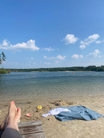 This is a horizontal, eye-level, outdoor photograph taken at a beach in Zheleznyi Perebor, Russia. The foreground shows a person's legs with pink toenail polish resting on a wooden structure. A blue blanket and a white cloth are spread out on the sand next to the wooden structure, along with a pink child's shovel and a yellow child's toy. The middle ground features a body of water with clear, shallow water near the shore and deeper, rippled water further out. The background consists of a dense, green forest bordering the lake, with a clear blue sky with scattered white clouds above. A power line is visible across the horizon, stretching from left to right. In the distance, on the water, a person can be seen on an inflatable raft.