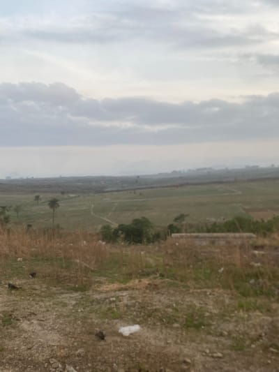 The image depicts a landscape in Lugbe, Nigeria. A field of mixed grasses and sporadic trees stretches into the distance, under a cloudy sky. The foreground shows a rough, uneven patch of ground with sparse vegetation and debris. There is no evidence of human presence or activity.