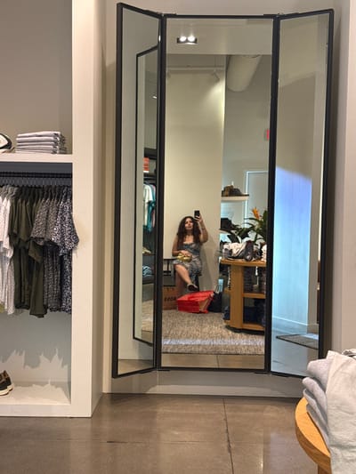A hinged mirror in a retail store reflects a woman taking a selfie with her phone. She is sitting on a box in the reflection. The store has clothing racks and displays. The floor is concrete. A table with folded clothes sits to the right of the mirror.