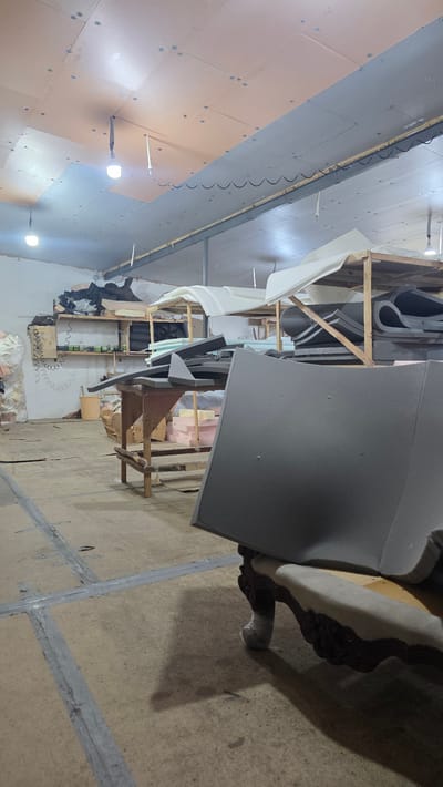 The image depicts an interior scene, likely a workshop or storage area, with a focus on various foam materials and furniture components. The setting appears to be a somewhat cluttered industrial space. In the foreground, a large piece of grey foam is prominently displayed, casting a shadow on the concrete floor. The floor itself has visible grey tape or sealant lines, suggesting construction or repair work.

To the left and in the background, wooden shelving units are filled with stacks of differently shaped and colored foam pieces, including white, grey, and pink. Some of these foam pieces are curved or irregular, suggesting they are intended for upholstery or cushioning. A partially visible piece of furniture, possibly a chair or sofa armrest with ornate dark wood detailing, is present in the upper right portion of the frame.

Further into the background, there are glimpses of electrical wiring, fluorescent lighting fixtures attached to the ceiling, and what appears to be a small person in dark clothing near the shelves. The overall lighting suggests an indoor environment, possibly during daylight hours or under artificial lighting, without any indication of weather conditions. There is no discernible text in the image. The scene evokes a sense of a workspace dedicated to crafting or preparing materials for furniture or insulation. The location context suggests this is in Novyy Chirkey, Russia.