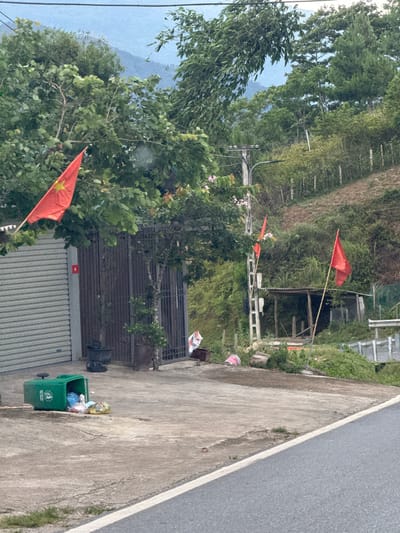 This image depicts a rural or semi-rural scene in Tu Le, Vietnam, characterized by lush green hills and a paved road. In the foreground, a gray asphalt road with a white solid line on its edge transitions to a concrete surface on the left. A green plastic wheelie bin lies on its side on the concrete, spilling multicolored plastic bags of trash. Adjacent to this, a building features a gray corrugated metal roll-down garage door and a dark metal gate. A large Vietnamese flag (red with a yellow star) is flown on a pole partially obscured by trees to the left of the building, and a smaller version of the flag is affixed to the building's wall next to the gate. Several potted plants are positioned near the gates.
Further to the right, a concrete utility pole with power lines and a streetlight fixture stands amidst dense foliage. Two plain red flags are erected on poles further up the sloped terrain. A rudimentary wooden structure with a corrugated metal roof is visible on the hillside behind a dirt path and a simple fence line. The background is dominated by steep, tree-covered mountains under a hazy sky. No individuals or animals are present.