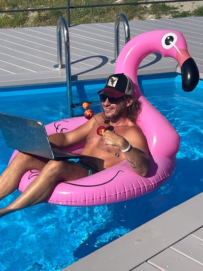 The image shows a person relaxing in a swimming pool on a pink flamingo float. The person is wearing a baseball cap and sunglasses, and appears to be working on a laptop. There are also two peaches on a skewer held by the person. The pool is surrounded by a wooden deck, and the scene appears to be set on a sunny day, given the bright lighting and shadows. The person is smiling, suggesting a relaxed and enjoyable mood. The location is identified as Мысовское сельское поселение, Ukraine.
