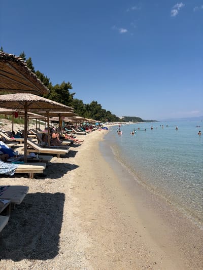 The image depicts a sunny day at a beach in Kassandra Municipal Unit, Greece. In the foreground, rows of lounge chairs with thatched umbrellas are set up on the sandy beach. Several people are seated on the lounge chairs, relaxing and sunbathing. The beach transitions into the clear, turquoise water of the sea, where numerous people are swimming and wading. Some individuals are using inflatable toys. The background shows a line of green trees and a distant building. The sky is a clear, bright blue with a few scattered white clouds. The overall scene conveys a sense of a typical summer day at a popular tourist destination.
