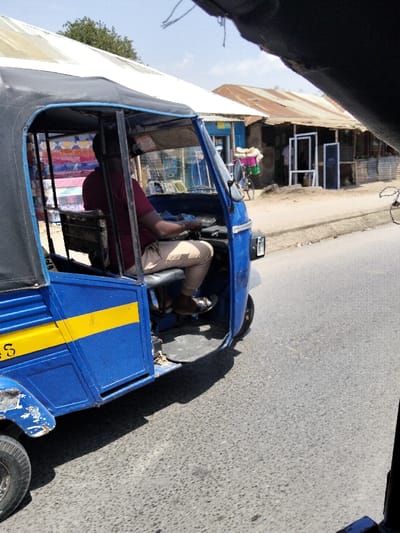 The scene depicts a blue and yellow three-wheeled auto-rickshaw (tuk-tuk) on an asphalt road in Mkomani ward, Kenya, under bright daylight. A male driver, with dark skin, wearing a maroon short-sleeved shirt and light-colored trousers, is seated in the driver's compartment, holding the handlebars. The tuk-tuk features a black fabric canopy and a horizontal yellow stripe on its blue exterior. Stacked items, resembling mattresses or textiles, are visible within the rear passenger section. To the right of the tuk-tuk, a dirt roadside borders the asphalt, leading to several single-story commercial structures. One building has a blue facade, while an adjacent structure features a weathered, rusty corrugated iron roof and displays multiple white window or door frames on its exterior. A bicycle laden with colorful goods is partially visible near these buildings. Green foliage and trees are present in the distant background. The photograph is taken from an elevated perspective, with portions of the camera's immediate foreground obscuring the upper edges of the frame.