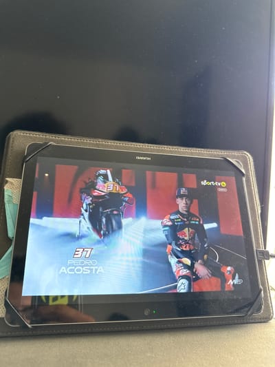 The image shows a tablet displaying a broadcast from Sport TV 4, Directo. On the screen, a motorcycle with the number 37 is visible, along with the text "PEDRO ACOSTA". To the right, a person in racing attire, also with the number 37 on their suit, is standing. The tablet is placed on a surface that appears to be a desk or table in what looks like a room, possibly a home or office. The background of the tablet screen features a red and black pattern. There is no indication of the city of Almada, Portugal in the image.