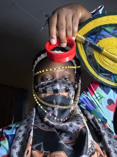 An individual with dark skin and multiple necklaces is partially obscured by a colorful, patterned headscarf. They wear a face mask and hold a red plastic ring near their face, with a woven hand fan featuring yellow and green details also in the frame. The scene appears to be indoors, with a dark floor visible in the background. It is not possible to determine the location of the image within the city of Madangala, Nigeria.