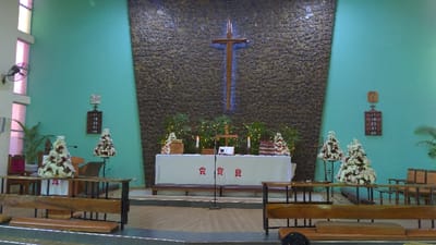 A church interior features a stone wall with a large cross behind the altar. The altar is covered with a white cloth adorned with red symbols. Two floral arrangements flank the altar, complemented by greenery and candles. Wooden pews face the altar, separated by railings. Wall-mounted date boards are visible on either side. The walls are painted a turquoise color.