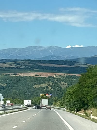 The image depicts a highway scene in Blatino, Bulgaria, on a clear day with blue skies and scattered clouds. A multi-lane highway curves to the right, bordered by a metal guardrail and reflective markers. Several vehicles are visible on the road, including a white truck with a trailer, a black car, and other trucks and cars further in the distance. The surrounding environment features rolling hills covered in lush green forests and fields, with a distant mountain range in the background. A large billboard is present on the right side of the highway. The perspective suggests the photo was taken from a moving vehicle.