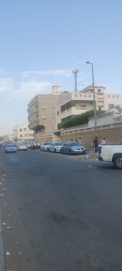 The image shows a street scene in Jeddah, Saudi Arabia, with several cars parked along the side of the road. Buildings line the street, including one with multiple air conditioning units. A telecommunications tower is visible on top of one building. Several people are standing on the sidewalk near the cars. The street is littered with debris. The sky is clear with some clouds.