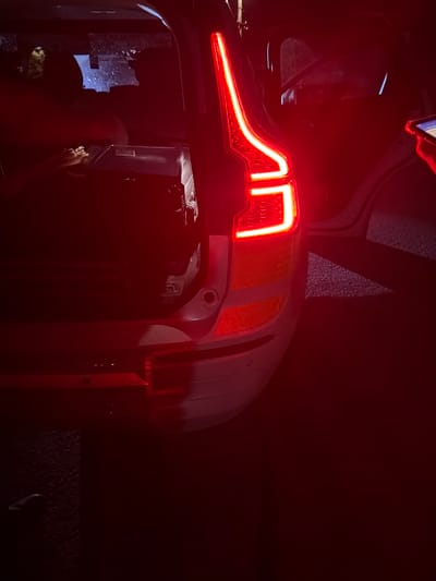 The image shows the rear of a white SUV with its tailgate open, illuminated by red taillights. Luggage is visible in the cargo area. The car is parked on a dark asphalt surface, possibly a street or parking lot, with some reflections of lights in the background. A hand holding a mobile phone is visible in the upper right corner. The scene is dimly lit, suggesting it is nighttime. The location is identified as Smyadovo, Bulgaria.
