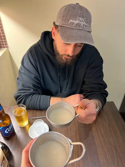 The image depicts two individuals seated at a dark brown wooden table within a casual restaurant setting in Chevilly-Larue, France, likely a Korean establishment.
One male subject, positioned on the right and facing downwards, is visible from the chest up. He wears a dark grey or black hooded sweatshirt and a distressed grey baseball cap with "C'EST magnifique" embroidered in white script on the front. He has a full beard. His right hand holds a silver-colored metal cup with a handle, containing a milky-white liquid. A silver-colored watch is visible on his left wrist, and a thin silver chain bracelet is on his right wrist. His left forearm rests on the table.
A second person, implied by a hand holding an identical silver-colored metal cup with a milky-white liquid, is positioned in the foreground to the left.
On the table between the two individuals are several items: a partially consumed bottle of "Cass" Korean beer, a clear glass filled with yellow liquid and foam (beer), a small white saucer holding a clear shot glass containing a clear liquid, a thin metallic straw or stirrer, and a dark-colored bottle with an indistinct label (possibly a condiment).
The background consists of plain light beige walls. A small section of reddish-brown square tiles is visible on the upper left wall. The lighting is artificial and warm.