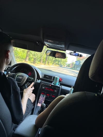 The image displays the interior of an Audi passenger vehicle, specifically a model identifiable by its steering wheel and dashboard design, during daylight hours in Lukovit, Bulgaria. The perspective is from the rear seat, looking forward.
A male driver, with short dark hair and wearing a black t-shirt, is positioned at the steering wheel, with his right hand resting on the lower spoke. The instrument cluster shows an ambient temperature of 30.5°C. A manual gear stick is visible between the front seats. A smartphone, displaying a navigation map with a route highlighted and indicating "32 min" travel time and "17:41" current time, is mounted on the center console. The car's audio system displays "101.7" on its screen. A light blue-green beverage can with a dragon motif is placed in a cup holder on the dashboard. A front passenger's arm and shoulder, covered by a light-colored garment, are partially visible on the right side of the frame.
The driver's eyes are reflected in the rearview mirror. Above the dashboard, a black dashcam is mounted on the windshield to the right of the rearview mirror, displaying a live view. The sun visors are in their stowed position against the dark gray headliner.
Outside the vehicle, a paved road stretches ahead, bordered by numerous trees with green foliage. Some tree trunks exhibit white bands. Buildings and utility poles are visible in the distance on the right side of the road. Other vehicles are also present on the road ahead. The scene is brightly lit by natural sunlight.