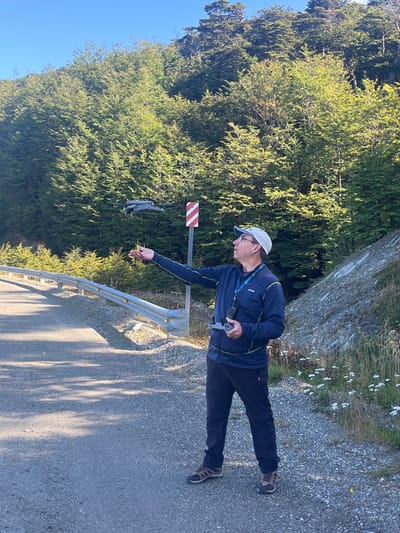 An outdoor scene in Lago Escondido, Argentina, depicts a male subject operating a drone. The man, appearing middle-aged, wears a light-colored baseball cap, glasses, a dark blue full-zip jacket with lighter trim, dark pants, and hiking shoes. A blue lanyard is visible around his neck. He stands on an asphalt road with a gravel shoulder, extending his right arm with an open palm towards a grey quadcopter drone that hovers in mid-air to his right. His left hand holds a drone remote controller. Behind him, a red and white striped marker post is visible. To the left, a metal guardrail with yellow reflectors follows the road's curve. The background is dominated by a dense forest of light green deciduous trees covering a steep hillside under a clear blue sky. To the man's right, a rocky embankment features scattered white flowers and dry vegetation. The scene is illuminated by direct sunlight.