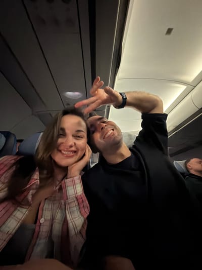 A photo shows two people, likely a couple, posing for a selfie inside an airplane cabin. A man with a black shirt, dark hair, and a wristwatch is partially visible at the top. A woman with long hair and wearing a pink and white plaid shirt is closer to the foreground and smiling. Part of another person is visible above the man. The background reveals parts of the airplane interior, including the windows, wall panels, and overhead storage. The location is likely the Емельяново (аэропорт) in Russia.