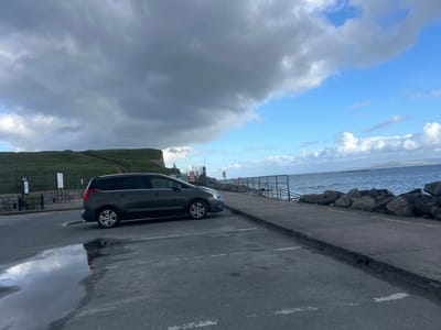 A grey multi-purpose vehicle is parked in a parking area adjacent to the coastline. To the left of the vehicle is a paved area with a puddle reflecting the sky. In the background, there is a grassy cliff leading down to the coastline. To the right of the vehicle, there is a concrete walkway, a metal railing, large rocks along the shoreline, and an expanse of ocean with land visible in the distance. The sky is a mix of dark, heavy clouds and patches of blue. The location is likely near Salthill Promenade, Galway, Ireland.